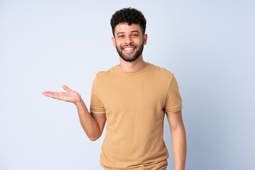 Young Moroccan man isolated on blue background holding copyspace imaginary on the palm to insert an...