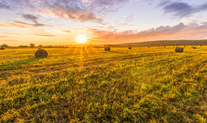Obraz premium Scenic view at picturesque burning sunset in a green shiny field with hay stacks, bright cloudy sky , trees and golden sun rays, summer valley landscape