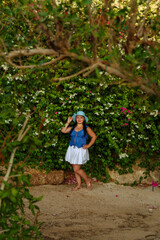 A brunette woman in a white skirt and a sun hat stands by the blooming white bougainvillea.