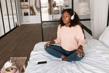 Relaxed African American Lady Meditating Wearing Headphones In Bedroom