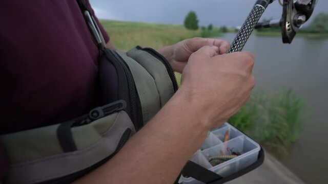 man fisherman puts on bait from fishing box hanging over his shoulder holding fishing rod in his hand in summer cloudy day close up