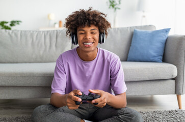 Portrait of cool black teen gamer with controller playing online video games, wearing headphones at...