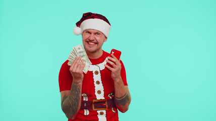Obraz premium Man in Christmas red t-shirt and hat looking mobile phone display sincerely rejoicing win, receiving money dollar cash banknotes, success lottery luck. Guy isolated on blue background. Happy New Year