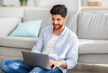 Young arab freelancer sitting with laptop on floor and leaning on sofa while working on computer at...