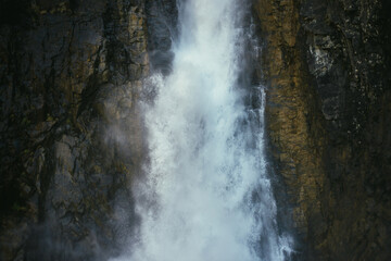 Atmospheric minimal landscape with vertical big waterfall on rock mountain wall. Powerful large waterfall in dark gorge. Nature background of high vertical turbulent falling water stream on wet rocks.