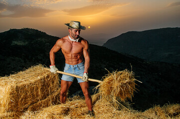Young shirtless farmer with sexy body, short jeans and hat with pitchfork in haystack.