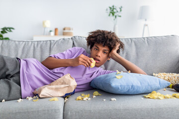 Bored Afro teenager lying on sofa with scattered snacks, eating chips, watching dull show or movie...