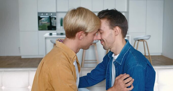 Portrait of two happy men gay couple in love kissing in make a gentle headbutt while sitting on sofa at home. LGBT Relationship