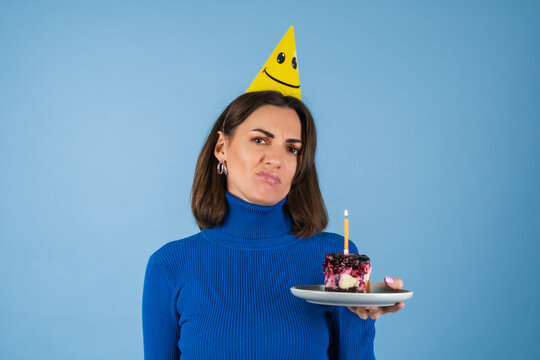 Young Woman In A Golf On A Blue Background Celebrates A Birthday, Holds A Piece Of Cake, Is Unhappy, Disappointed, Does Not Want To Grow Old, Lonely