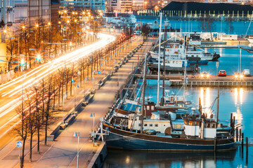 Fototapeta na wymiar Helsinki, Finland. View Of Pohjoisranta Street And Ships, Boats And Yachts Moored Near Pier In Evening Night Illuminations