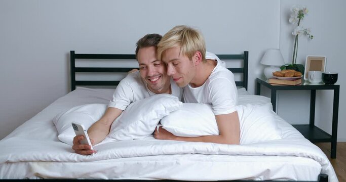 Homosexual couple hugging looking at smartphone camera when making selfie photos. Two men gays lying on bed in bedroom and taking photos with mobile phone
