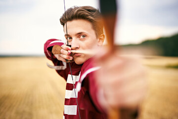 Young male sportsman targeting with bow in a traditional, medieval archer costume - Teenager archer practicing archery in nature - Field archery and recreation concept with a millennial boy - obrazy, fototapety, plakaty