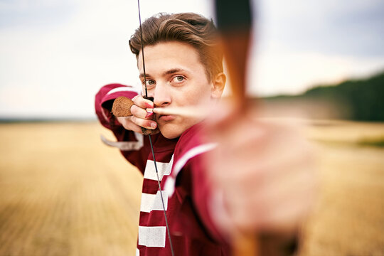 Young male sportsman targeting with bow in a traditional, medieval archer costume - Teenager archer practicing archery in nature - Field archery and recreation concept with a millennial boy