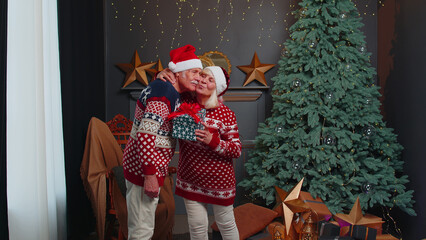 Happy elderly couple in Santa Claus hats celebrating New Year in cozy living room at home, grandfather gifting Christmas present box to surprised grandmother. Mature people celebrate Christmas at home