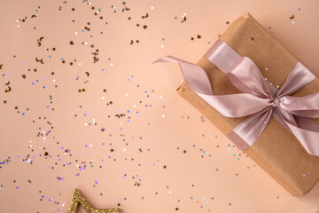 Birthday or New Year's eve present in kraft paper with pink ribbon on a soft beige background with glitter and confetti. Xmas composition. Flat lay. Happy holidays celebration and giving love concept