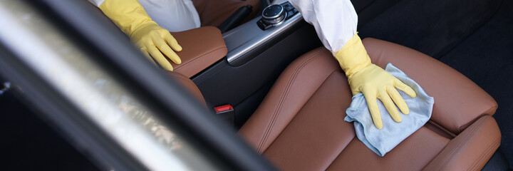Man in protective suit and rubber gloves wiping leather car seat with rag with disinfectant closeup