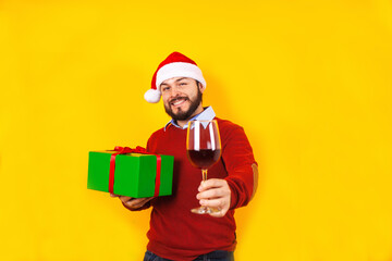 latin man in christmas concept holding a glass of wine with santa hat, having fun, smiling amazed on a yellow background in Mexico Latin America