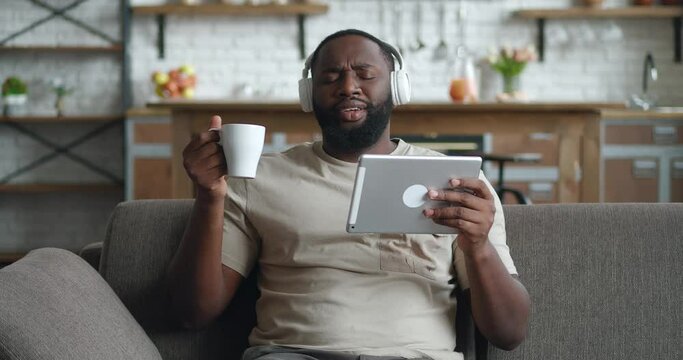 Smiling black man in headphones using digital tablet sit on couch at home, young african guy drinking coffee holding computer looking at screen reading e-book app listening music online