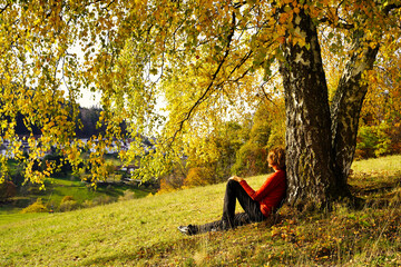 A caucasian woman sitting under a birch tree with yellow autumn leaves. The sun is shining. A few houses from a village in the background. Germany, Baden Wurttemberg, Wilhelmsfeld.
