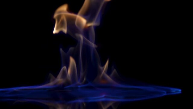 Falling match causes flames to ignite against black studio background. Combustion flammable liquid close up. Spot of gasoline ignites with flash and burns with blue yellow light in dark in slow motion