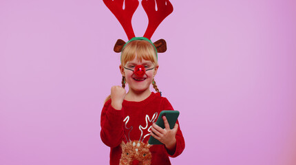 Girl toddler in Christmas red sweater deer antlers looking smartphone display sincerely rejoicing...