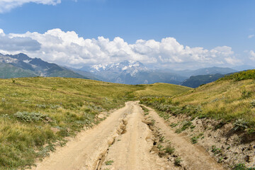 Fototapeta na wymiar Summer mountain landscape with a dusty road in Svaneti region, Georgia, Asia. Snowcapped mountains in the background. Blue sky with clouds above. Georgian travel destination