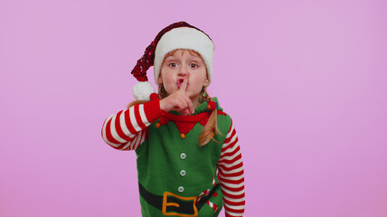 Keep my secret. Do not tell anyone. Shh be quiet please. Child girl kid in Christmas Santa Claus helper Elf costume presses index finger to lips makes silence gesture sign. Happy New Year celebration