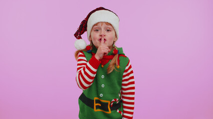 Shh. Keep my secret. Do not tell anyone. Quiet, please. Child girl kid in Christmas Elf Santa Claus helper costume presses index finger to lips makes silence gesture sign. Happy New Year celebration