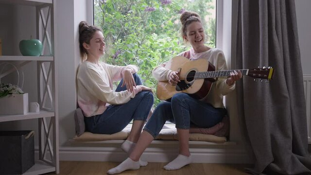 Wide shot talented beautiful twin sisters sitting on windowsill playing guitar singing. Portrait of confident Caucasian charming identical women enjoying hobby indoors at home on weekend