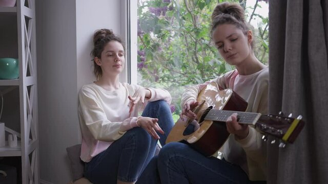 Portrait of relaxed happy beautiful woman singing with twin sister playing guitar sitting on windowsill at home. Carefree Caucasian slim women enjoying hobby on weekend indoors