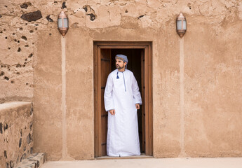omani man in traditional outfit at a doorway of an old house - obrazy, fototapety, plakaty