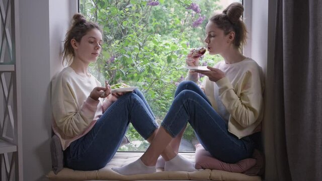 Slim beautiful identical twins eating healthful salad and unhealthy sweet bun. Wide shot side view Caucasian young women with different food sitting on windowsill at home at lunch