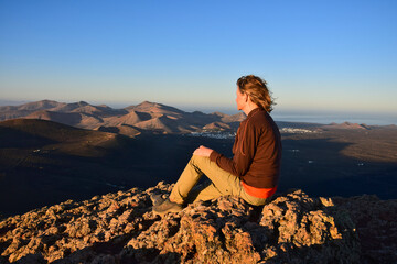 Sunrise in Lanzarote. A woman sitting on a mountain, overlooking the volcanic landscape with the small towns Yaiza and Uga. Canary Islands, Spain.