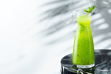 green summer lemonade in jug on light background with shadow of branch tropic plant. Lemonade with kiwi and ice