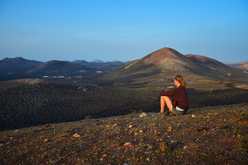 Sunrise in Lanzarote. A woman sitting on a mountain, overlooking the landscape of La Geria and Timanfaya National Park. Canary Islands, Spain.