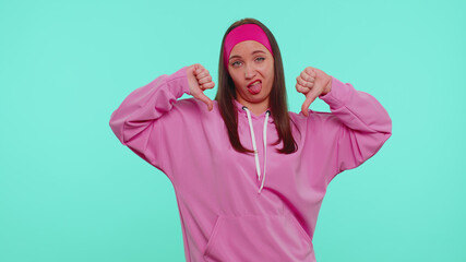 Fototapeta premium Upset pretty stylish teen girl in pink hoodie showing thumbs down sign gesture, expressing discontent, disapproval, dissatisfied, dislike. Young adult woman. Indoor studio shot on blue wall background