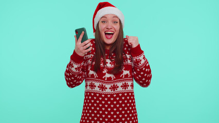 Wow. Girl in stylish Christmas red sweater and Santa hat looking smartphone display sincerely rejoicing win, receiving message with good news, success luck. Woman on blue background. Happy New Year