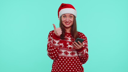 Wow, super. Girl in Christmas sweater looking smartphone display sincerely rejoicing win, receiving message with good news, success luck, showing thumbs up. Woman on blue background. Happy New Year