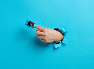 an oximeter for measuring oxygen in the blood is worn on the index finger, part of the hand is sticking out of a torn hole in the blue paper - obrazy, fototapety, plakaty
