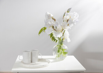 A bouquet of three white irises and a fern in a transparent vase on the table. Two ceramic tea cup in the tray. Breakfast