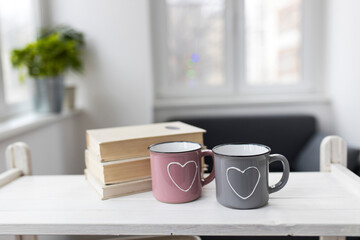 Fototapeta na wymiar Gray and pink with hearts enamel cups sit on the table in front of the window. Valentine's Day.