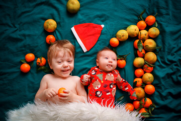 The older brother lies with a Christmas newborn baby on a cozy green bed, next to Santa's hat and the number one of the tangerines. The babies are covered with white fur. High quality photo