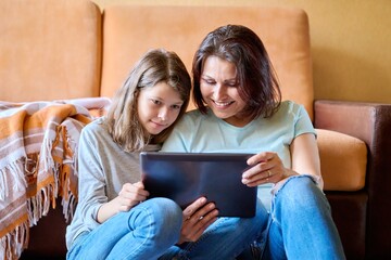 Middle-aged mom and preteen daughter relaxing at home together, with digital tablet