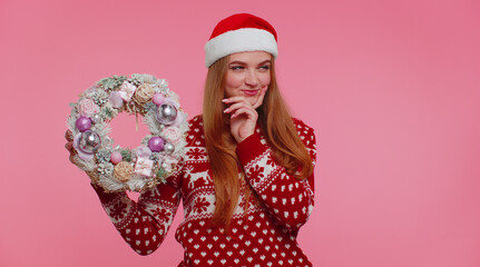 Thoughtful one adult girl 20s in red Christmas deer sweater look camera smiling hold raise point finger on festive wreath isolated on light pink background studio. Happy New Year holiday celebration