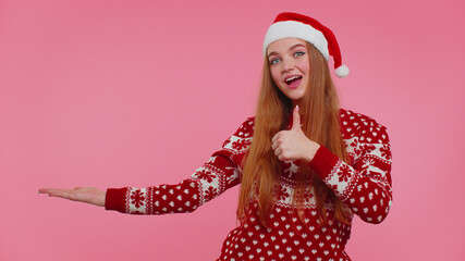 Adult teen cute girl wears red Christmas deer sweater showing thumbs up and pointing at left on blank space. Place for your advertisement logo on red background studio. Happy New Year holidays sale