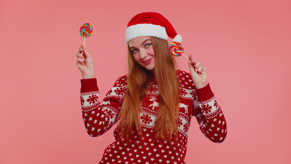 Funny girl in red Christmas sweater, hat holding candy striped lollipops, hiding behind them, fooling around, dancing isolated on pink studio wall background. Happy New Year celebration Xmas holidays