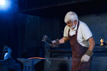 Professional blacksmith with grey beard and hair beating molten steel with heavy hammer. Caucasian man in safety apron and gloves hardening iron on anvil at forge.