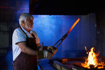 Fototapeta premium Caucasian craftsman with grey beard and hair preparing metal for shaping while heating it in burning fire. Concept of people and blacksmithing. 