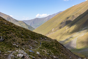 A female backpacker on a hiking trail with a panoramic view on the green hills and sharp ridges of the mountain peaks in the Greater Caucasus Mountain Range in Georgia, Kazbegi Region. Wanderlust.