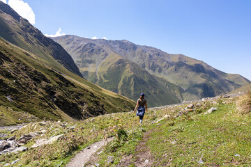 Fototapeta na wymiar A female backpacker on a hiking trail with a panoramic view on the green hills and sharp ridges of the mountain peaks in the Greater Caucasus Mountain Range in Georgia, Kazbegi Region. Wanderlust.